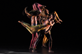 A cluster of dancers lifting and supporting eachother in front of a black background.